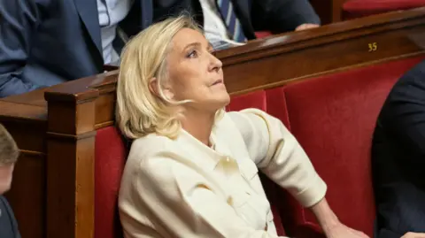 Marine Le Pen sits in France's National Assembly in a red velvet seat. She is biting her lip and looking up. She wears a cream suit. 
