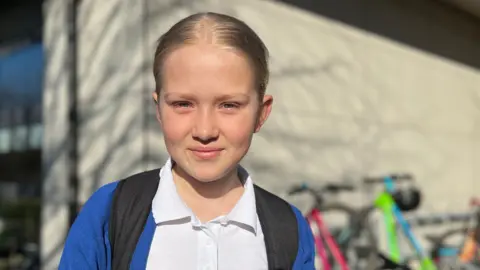 Dorota is wearing a white polo shirt, blue cardigan, and a backpack stands outdoors near a school building. Several bicycles are parked in the background on a sunny day.