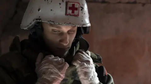 Lee Durant/BBC Denys wearing a white helmet with a red cross on it. The atmosphere is sombre and he's looking pensive.