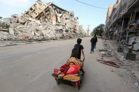 Reuters A Palestinian boy pulls a cart carrying his brother and their belongings as they flee their home during Israeli air and artillery strikes, near the site of destroyed a tower block in Gaza City (14 May 2021)