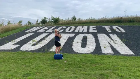 Max's Missions/Facebook Georgie Dunt with her cat Max, standing next to a sign which says Welcome To Luton