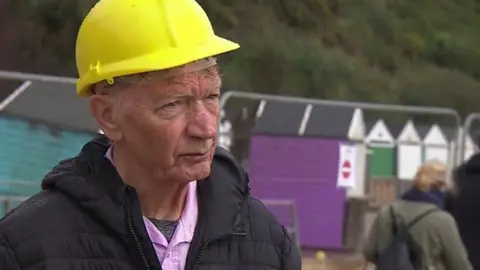 Man wearing yellow hard hat, pink shirt and black coast metal fencing and purple and turquoise beach huts are behind him.