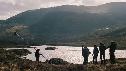 Black Camel Pictures/BBC Alba/John Maher Filming taking place on the Isle of Harris - members of the crew are standing with cameras and equipment, near water that has the sun shimmering down on it. The hills and greenery can be seen in the distance and around them.