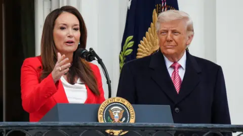 EPA/Shutterstock US Secretary of Agriculture Brooke Rollins wears a red blazer and peaks as President Donald Trump listens during an event celebrating farmers and Agriculture Day on the South Lawn of the White House in Washington, DC, on 27 March 2026.