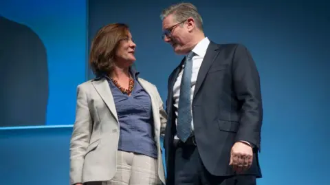 Eluned Morgan stood alongside Sir Keir Starmer at Welsh Labour conference last year. Morgan is wearing a blue top and grey suit, while Sir Keir is wearing a black suit, a blue tie, a white shirt and glasses.