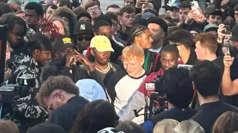 A ginger-haired man wearing a white t-shirt with black markings is surrounded by people.