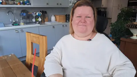 BBC Jennifer Gallacher is sitting smiling at her kitchen table wearing a white jumper. There is a wooden chair behind her as well as grey kitchen units with a knife block and mugs on a stand. 