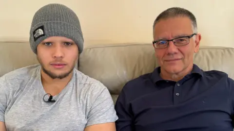 Charlie Rose/BBC In a grey t-shirt and beanie hat, George sits on a beige leather sofa next to his dad Tom who is wearing a navy top and glasses. Father and son are looking directly at the camera.
