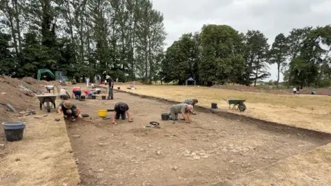 Josh Gorrono-Chapman/BBC A group of people on their hands and knees on a large plot of soil. They are picking up bits of Roman material that has been discovered underneath the ground.