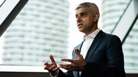 Side profile of the mayor of London Sadiq Khan being inverviewed. He is wearing a dark suit and a white shirt and is gesturing with his hands. There are tall buildings in the background.  