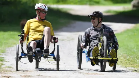 Two men riding recumbent bikes, wearing cycle helmets and laughing to each other 