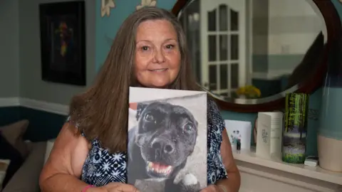 Fiona stands indoors holding a printed photograph of a black dog close to her chest. The print shows a close-up of the dog’s face, with one eye prominently visible and the mouth slightly open. Fiona has long hair and is wearing a patterned top, along with a pink wristband on one wrist.
In the background, a softly lit room is visible with teal-coloured walls, a round mirror mounted on the wall, and a white shelf displaying a card with a paw print and small decorative items, including a tall, green-toned glass ornament.