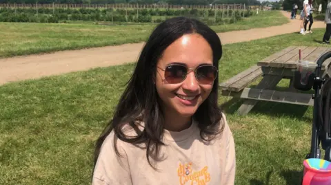 ALEXANDER STEVANOVIC/BBC Danielle is sat on a bench.
She is wearing shades and a cream coloured jumper and has dark hair.
In the background is the fruit & veg farm.