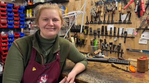 Lucy Cushely has blonde hair pulled back behind her head and is wearing a green polo kneck, green sleveless fleece and a maroon apron. She is sitting in her workshop, her left elbow rests on a work bench and behind her is an array of leather-working tools.