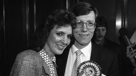 Pacemaker Iris and Peter in a black and white photo in 1987. Iris has long dark hair and Peter has short dark hair and wearing glasses.