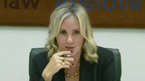Dame Caroline Dinenage chairs a meeting in parliament. She has wavy blonde hair and wears a dark jacket and a gold chain necklace.