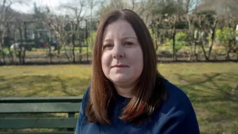 Lucy's head. It has long, dark and straight hair and wears a blue blue shirt. It was filmed abroad, on a green bench, with bushes behind it.