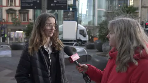 Nia Price, who has blonde long hair and i wearing a red jacket, holding a microphone out in front of a girl with long brown hair and glasses wearing a black jacket. They are on a pedestrianised street with buildings, benches and delivery vehicles in the background