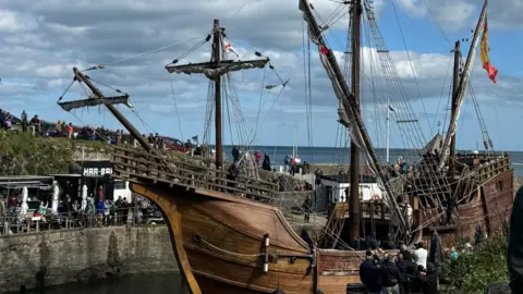 A tall ship in a harbour. There is a Spanish flag. There are crowds of people watching the ship. The sky is blue and cloudy.