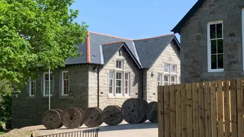 A view through trees and a wooden fence to a granite bunkhouse building. There is a row of wooden spools in front of it.