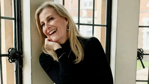 Sophie Wessex, who has long blonde hair and wears a black turtleneck, laughs in a photo in which she poses by a bay window with her hand cupping her face