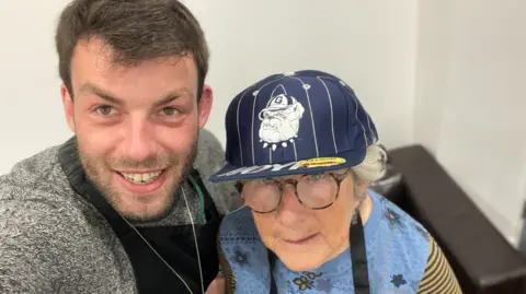 Zink A man with brown hair and beard smiling at the camera stood next to an elderly lady in a cap