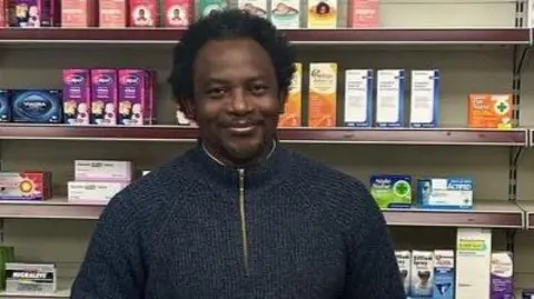 Tunde Olawoye stood behind the counter at his pharmacy wearing a blue fleece jumper. There are shelves of drugs and medicine behind him.