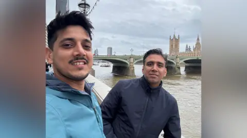 Jitender Tarar Two men stand along what appears to be the Thames, with London Bridge behind them. The man on the left has short black hair, a short beard and is wearing a light blue coat. The man on the right has short black hair that is greying, and is wearing a dark black coat.