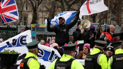 Reuters A man with a black beard, baseball cap, black top and sunglasses waves and Israel flag stands above a crowd of protesters. A man to his left also dressed all in black is speaking into a megaphone. The protesters are waving UK flags and Israel flags. They are in front of the Belfast Christmas market, the top of stalls can be seen in the background with some fairly lights on them. Police stand in front of the protesters in the foreground. 
