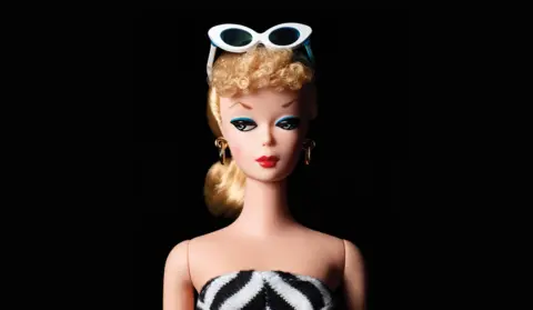 Studio portrait of a blonde fashion doll with short curly hair, wearing white sunglasses on top of the head, gold hoop earrings, bold red lips, blue eye makeup, and a black‑and‑white striped strapless outfit, set against a dark background.