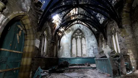 Geoff Hughes Inside of the chapel with broken bench on the right and rubble on the floor, with light peeking through gaps in the roof.