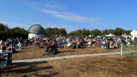 A picture of crowds gathering in a field. They are sat on a grass field.