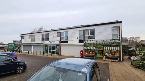 A white fronted building with grey horizontal shutters. A fruit and vegetable  shop is open.