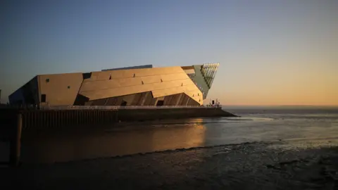 A view of The Deep. It is a silver metal-clad triangular building with a glass point at the front. It stands on the side of the Humber Estuary and the River Hull and has sun reflecting off it.