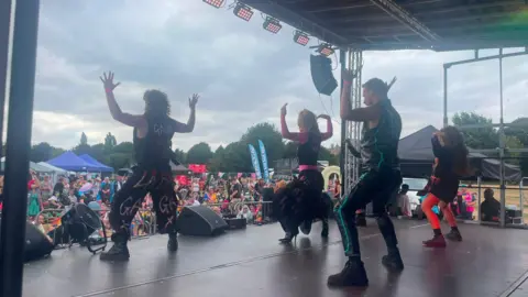 Peterborough Pride Four people are on a stage dancing. There are crowds of people watching and milling around and there are tents and stalls in the background.