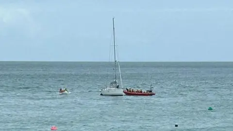 Jersey RNLI A small white yacht in blue seas. It is surrounded by two lifeboats. There are a couple of bouys dotted in the distance. The sky is blue. 