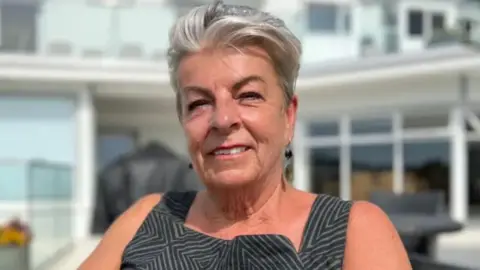 Penny Bromley smiles while sitting in a chair in a garden. She is wearing a black and grey dress and has black dangly ear-rings.
