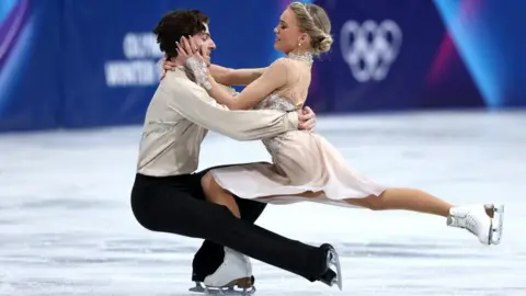 Two figure skaters in each other's arms on an ice rink. They are dressed in dancing outfits are are in the middle of their routine.