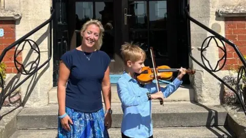 Pro Corda Annabel Thwaite with her son outside Framlingham College. She has blonde hair and is wearing a blue T-shirt and skirt and is smiling. Her son is playing the violin