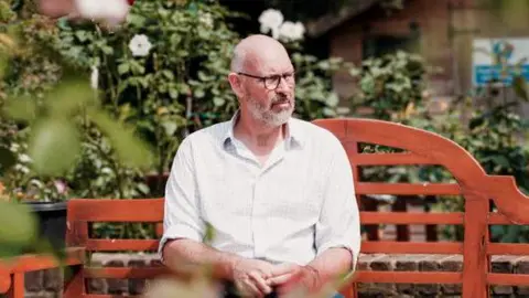 National Lottery/PA man with glasses sits on a bench surrounded by shrubbery