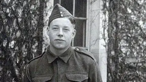 A black and white photograph of James Conway wearing military uniform and hat. He is stood in front of a white building, which has plants growing up the walls and is looking at the camera.