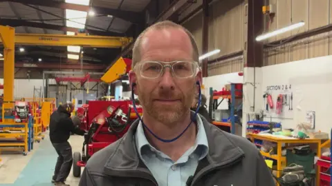 A man stands in a factory assembly area , behind him a man puts the final touches to a large piece of farm machinery. Tom wears protective glasses and ear plugs 