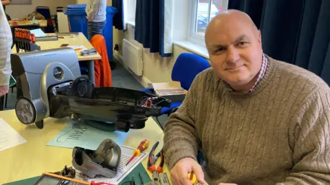 Paul Bramall, in a brown knitted jumper, is working on the repair of a vacuum cleaner