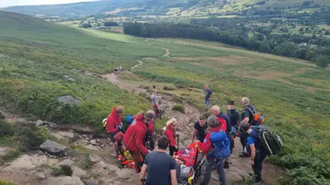 A team of volunteers in outdoor clothing carry a person a stretcher down a path towards trees at the bottom of a valley