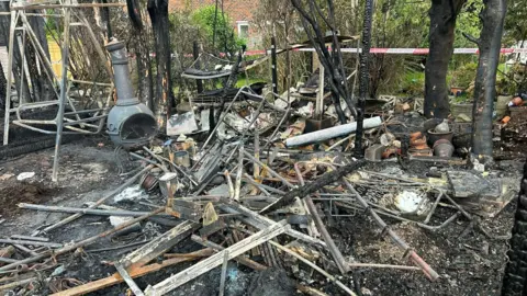 A pile of burned wreckage sat on a charred surface in a garden. The remains of some trees, a garden swing and a garden stove can be seen amongst the ruins.