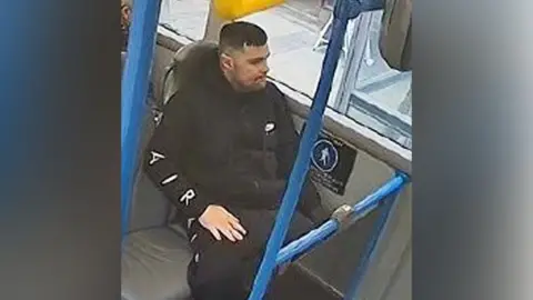 A man in a black tracksuit sitting on a bus, staring out of the window. The image is low quality and is taken from CCTV.