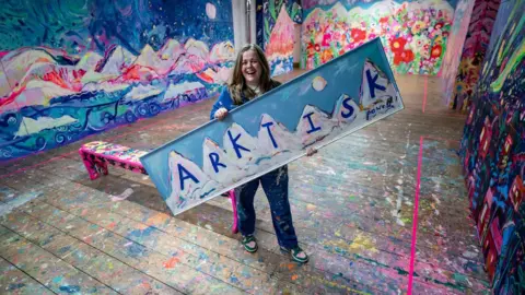 PA Media Emily Powell pictured holding a large sign reading "Arktisk" with mountains painted on it. 