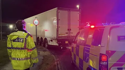 A police officer in a yellow jacked stood by a white/grey HGV pulled over by a police vehicle.