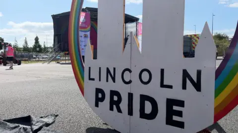 A colourful sign of Lincoln Pride's Logo (a silhouette of Lincoln Cathedral surrounded by a rainbow). In the background, workers are setting up a stage under a blue sky.