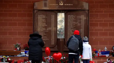 A woman and a man with a child stand in front of the memorial at Anfield that lists the names of those who died. Red heart shaped balloons, flowers, candles and picture frames are beneat the plaque of 97 names.